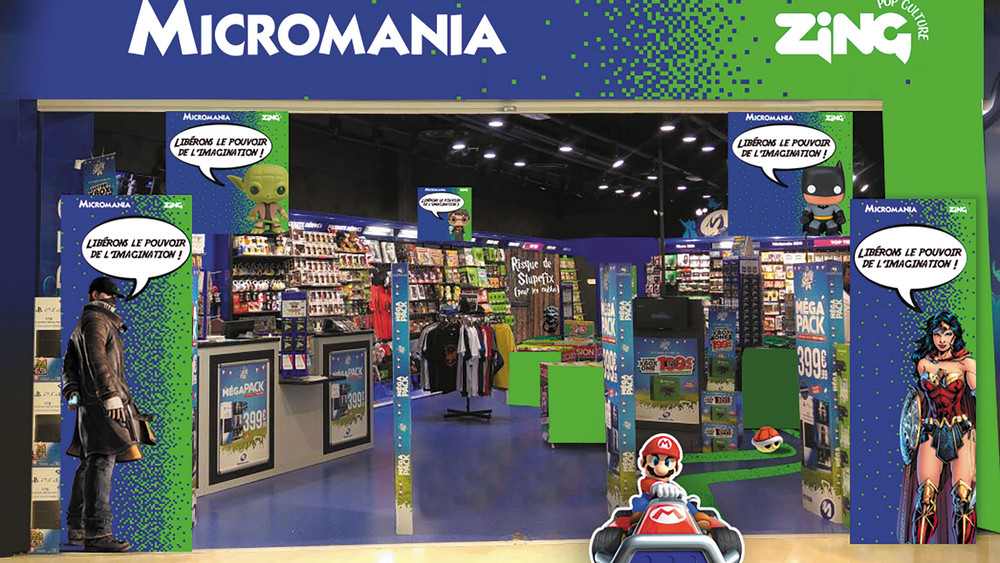 Visuel d’une boutique Micromania Zing avec une devanture bleue et verte, des rayons remplis de jeux vidéo, figurines et produits dérivés. Des illustrations de Mario, Wonder Woman, Batman, Groot et d’autres personnages décorent l’entrée, accompagnées du slogan « Libérons le pouvoir de l’imagination ! » et d’affiches promotionnelles pour des offres en magasin.