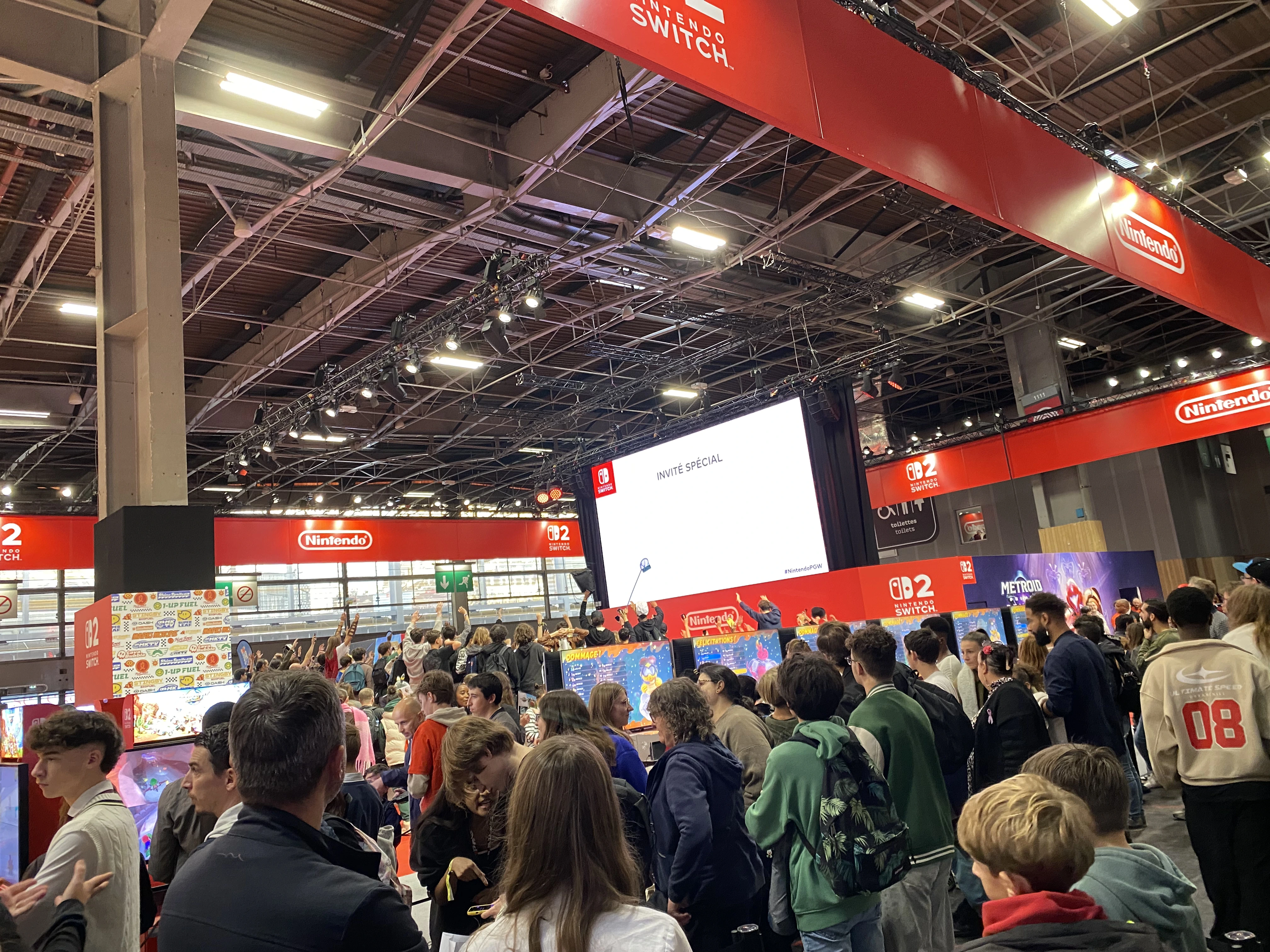 Vue large et animée du stand Nintendo à la Paris Games Week 2025, fortement fréquenté par les visiteurs. Sous une imposante structure rouge marquée du logo Nintendo et des mentions « Switch 02 », une foule dense se presse autour de plusieurs bornes de jeu. Un grand écran central indique « INVITÉ SPÉCIAL » en lettres noires sur fond blanc, laissant entendre la venue d’une personnalité. Plusieurs jeux sont mis en avant, dont Super Mario Bros. Wonder et Metroid Dread, visibles sur les affiches colorées. Les visiteurs, majoritairement jeunes, sont debout, certains tenant des sacs ou des objets promotionnels, et d’autres pointant leurs téléphones vers la scène. L’ambiance est festive et dynamique, typique des grands événements vidéoludiques. L’éclairage puissant du plafond met en valeur la scène et les décors aux couleurs vives de Nintendo.
