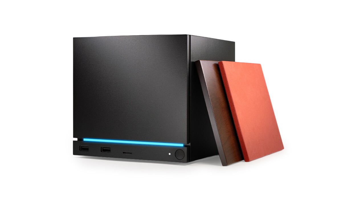 Visuel produit montrant un mini PC ou boîtier compact noir au design cubique avec une bande lumineuse bleue en façade, plusieurs ports de connexion et un bouton d’alimentation. Deux panneaux latéraux interchangeables, l’un effet bois foncé et l’autre rouge mat, sont appuyés contre l’appareil sur fond blanc, mettant en avant les options de personnalisation du châssis.