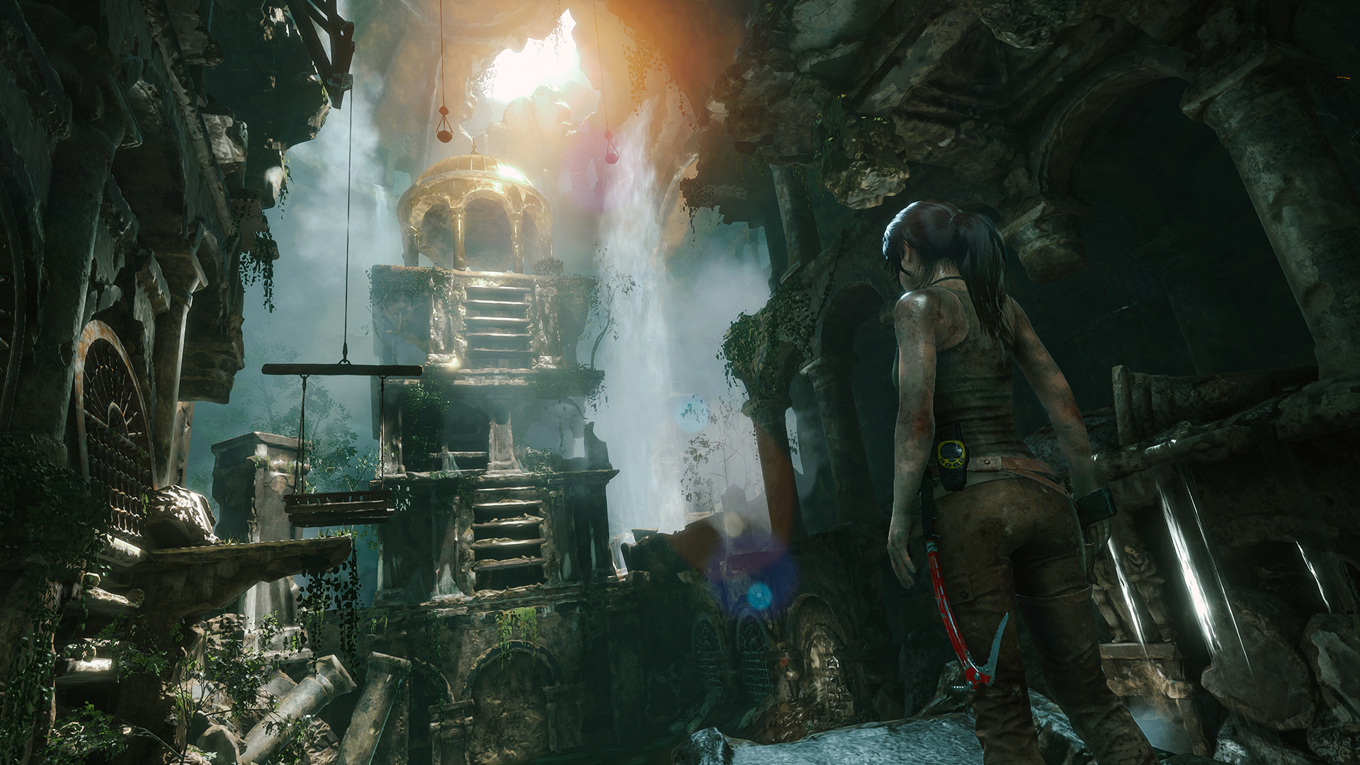 Capture d’écran de Rise of the Tomb Raider montrant Lara Croft observant des ruines anciennes au cœur d’une grande caverne. Des cascades, des plateformes de pierre, des plantes et une lumière venant du plafond créent une ambiance d’exploration mystérieuse et spectaculaire.
