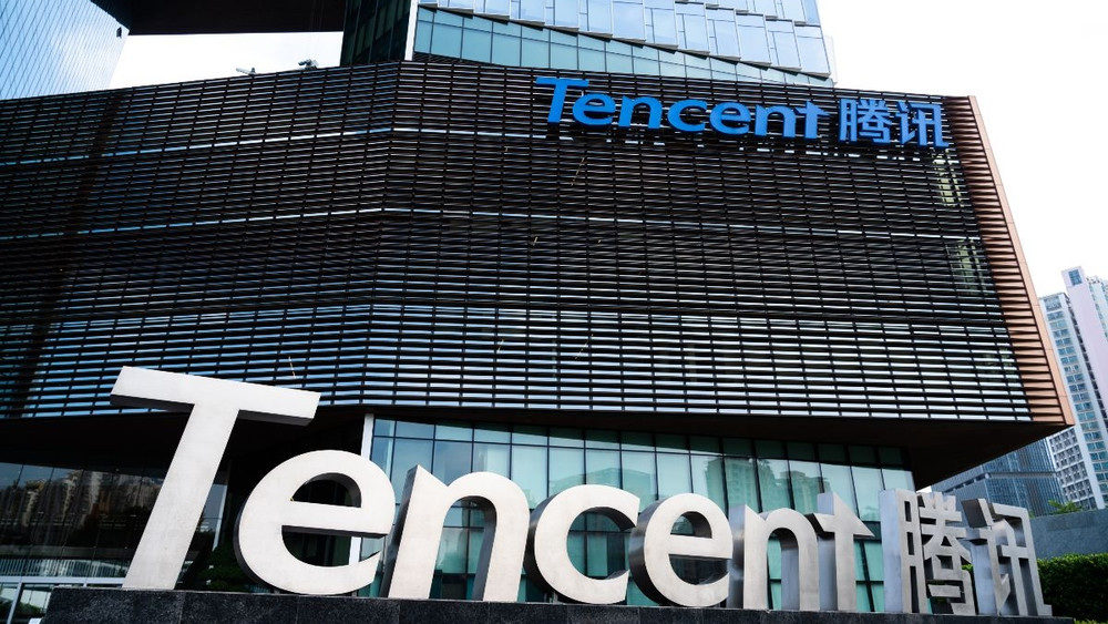 Vue extérieure du siège de Tencent, un immeuble de bureaux moderne en verre et métal à façade ajourée. De grands logos “Tencent” et “腾讯” apparaissent en relief sur le bâtiment, en bas et en haut, dans un environnement urbain de gratte-ciels.