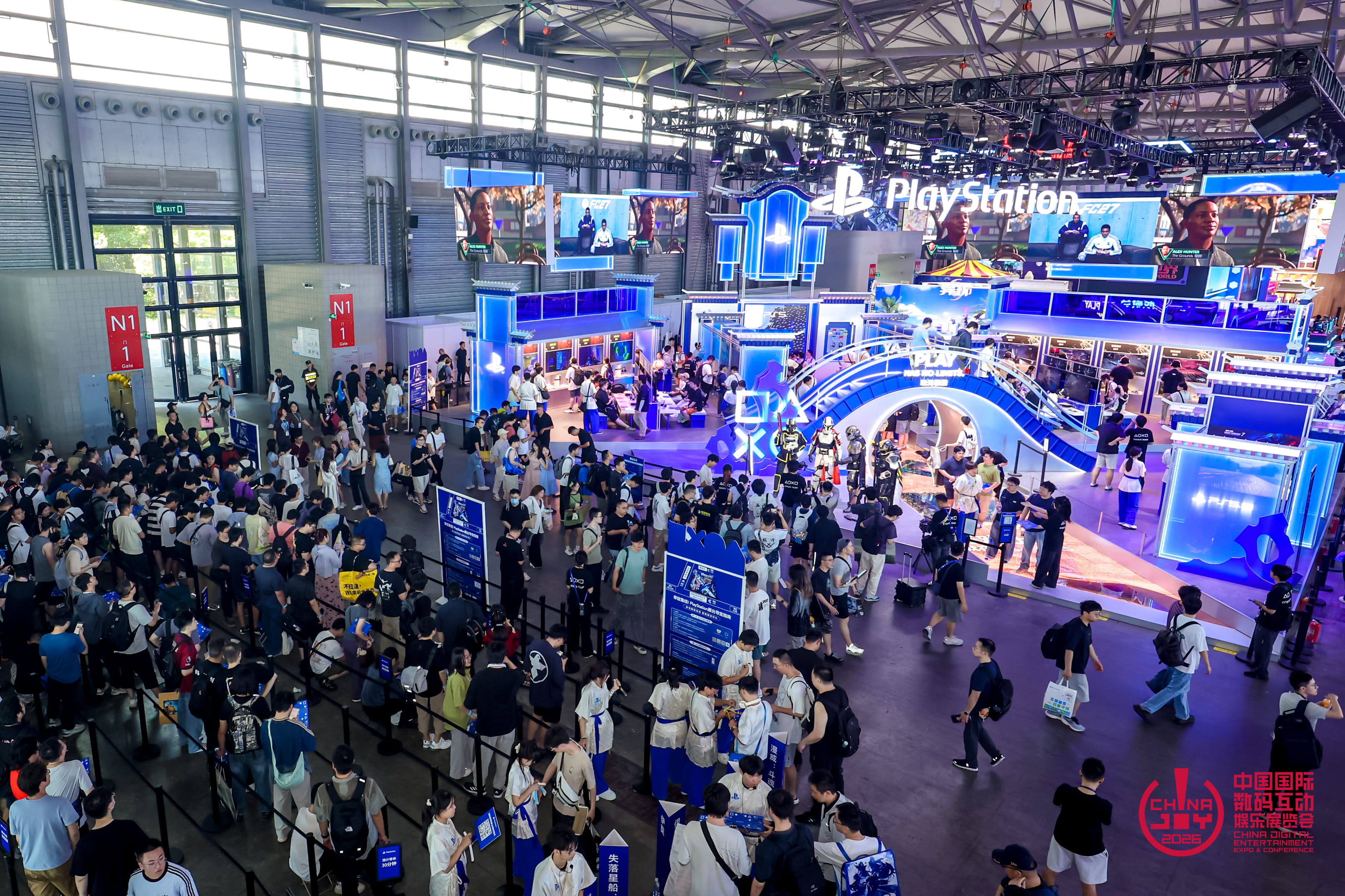 Vue en hauteur du stand PlayStation à ChinaJoy 2026, dominé par une grande enseigne lumineuse et des écrans géants. De nombreux visiteurs font la queue autour des espaces de démonstration bleus et blancs, avec plusieurs statues de personnages exposées au centre.