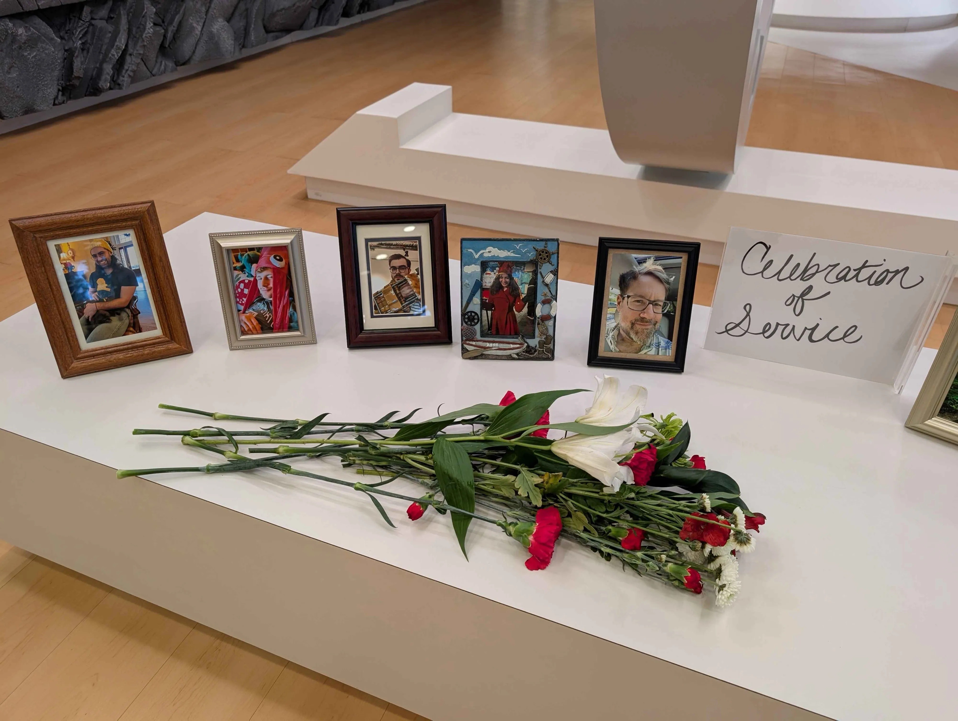 Photographie d’un mémorial installé sur une table blanche, composé de plusieurs photos encadrées de personnes, d’un bouquet de fleurs blanches et rouges et d’un panneau portant le texte « Celebration of Service ». L’ensemble évoque un hommage ou une cérémonie de commémoration dans un espace moderne aux lignes épurées.