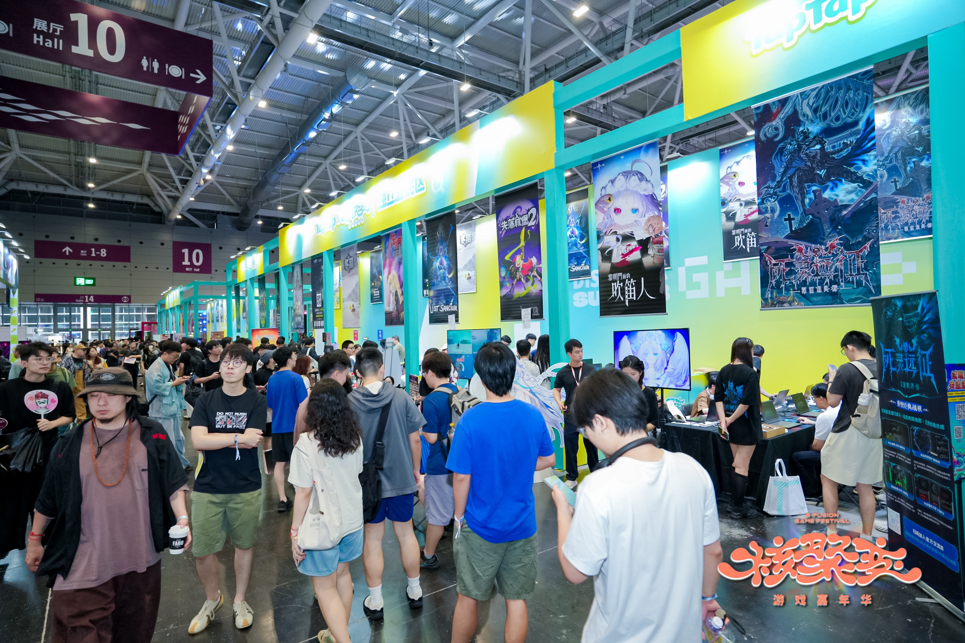Photographie d’une allée très fréquentée d’un salon du jeu vidéo dans le Hall 10 d’un centre d’exposition. De nombreux visiteurs circulent entre des stands colorés présentant des jeux indépendants, avec de grandes affiches illustrées et des écrans de démonstration. Les enseignes et panneaux comportent principalement des textes en chinois, tandis que le logo du festival apparaît en bas à droite de l’image.