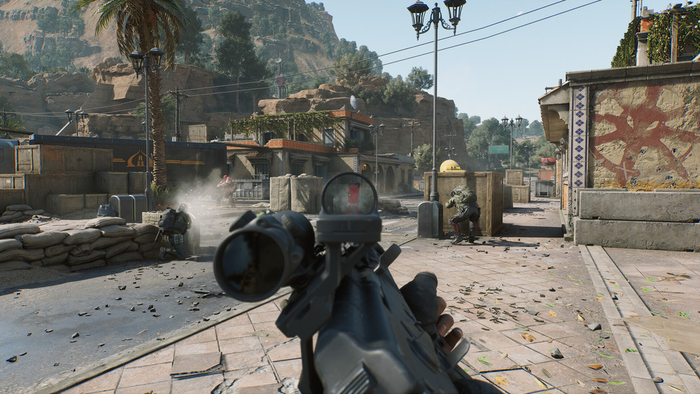 Capture d’écran d’une séquence de gameplay en vue subjective (FPS) tirée d’un jeu de tir militaire réaliste. Le joueur, visible par ses mains tenant un fusil d’assaut équipé d’une lunette holographique, vise un ennemi au loin, au centre du viseur. L’action se déroule dans un environnement urbain désertique, bordé de montagnes, de palmiers et de bâtiments en pierre inspirés d’une architecture moyen-orientale. Plusieurs soldats lourdement armés sont engagés dans un échange de tirs : un ennemi est touché en pleine rue, projetant de la poussière et des éclats ; un autre est accroupi derrière une caisse en bois sur la droite ; un troisième est à couvert derrière des sacs de sable à gauche. Les détails graphiques sont très fins, avec des effets réalistes de lumière, de particules et de textures, indiquant un moteur de jeu moderne. La scène illustre un moment de tension intense typique des jeux de tir tactiques multijoueurs comme Call of Duty: Modern Warfare II ou Warzone.