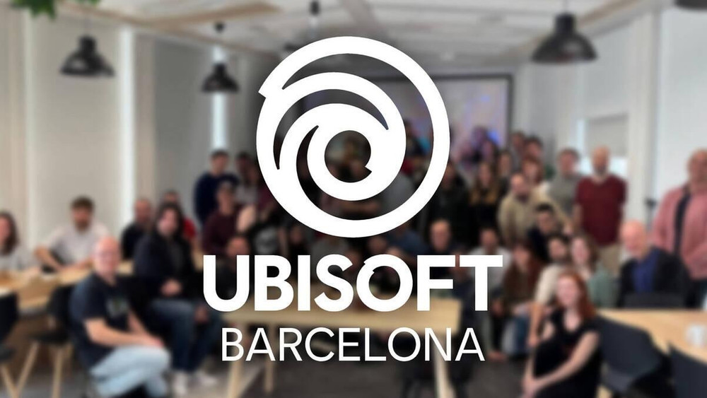 Logo Ubisoft Barcelona en blanc superposé à une photo de groupe floue montrant les équipes du studio réunies dans un espace de bureaux lumineux avec tables, chaises et suspensions au plafond. Aucun autre texte n’est visible en dehors du nom du studio.