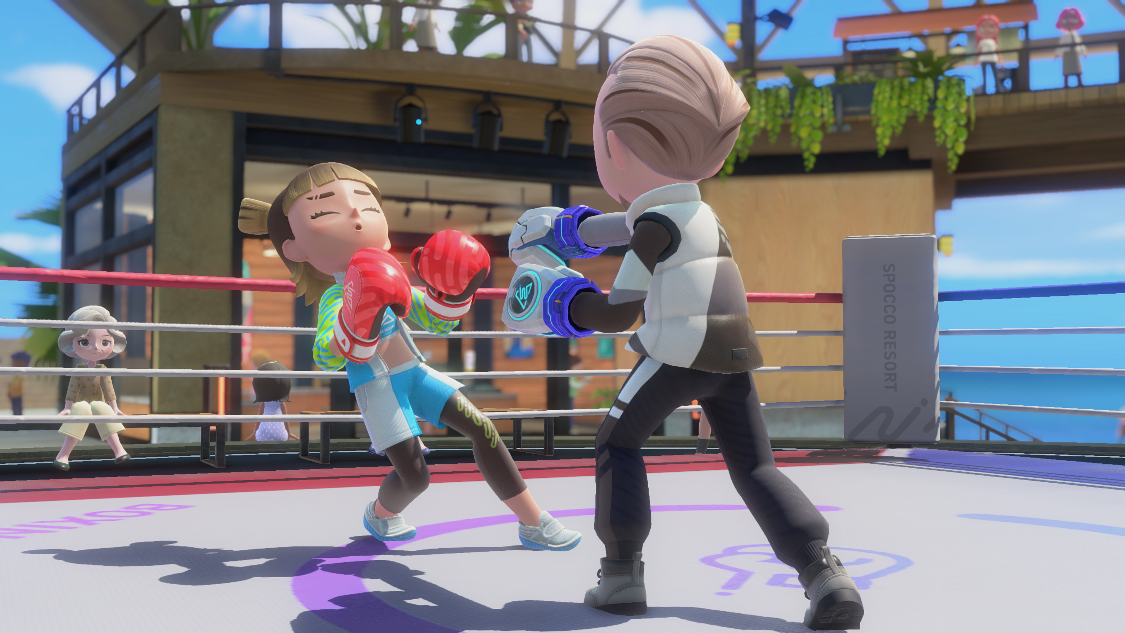 Capture d’écran de Nintendo Switch Sports Resort montrant deux personnages en plein match de boxe sur un ring en extérieur. L’un reçoit un coup avec des gants rouges tandis que l’autre attaque avec des gants bleus, sous les yeux de spectateurs dans une ambiance sportive et ensoleillée.