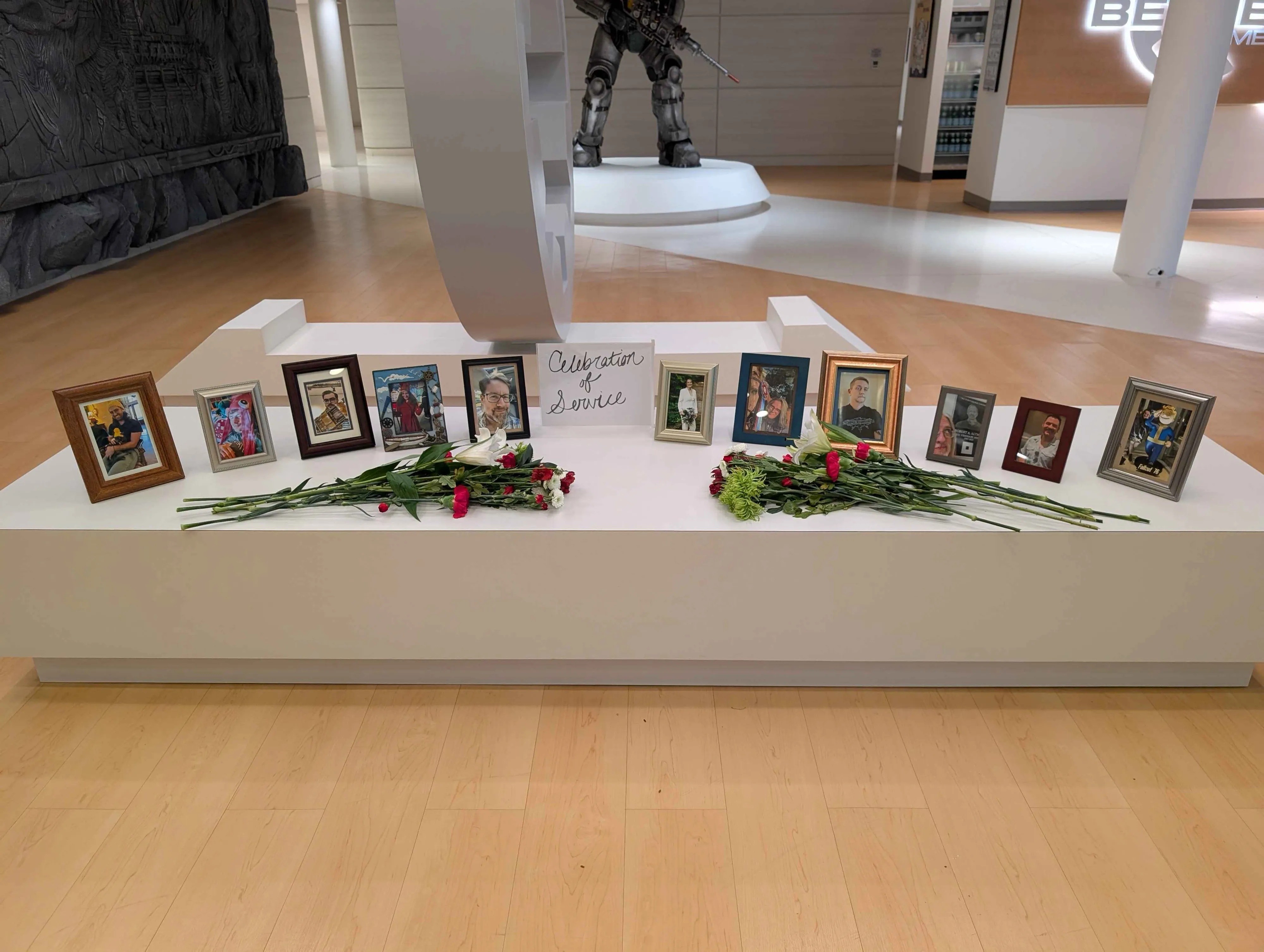 Photographie d’un mémorial installé dans un hall moderne, composé de plusieurs photos encadrées alignées de part et d’autre d’un panneau portant le texte « Celebration of Service », avec deux bouquets de fleurs blanches, rouges et vertes déposés devant. En arrière-plan, une grande sculpture blanche et une imposante statue en armure évoquent un siège d’entreprise lié au jeu vidéo.
