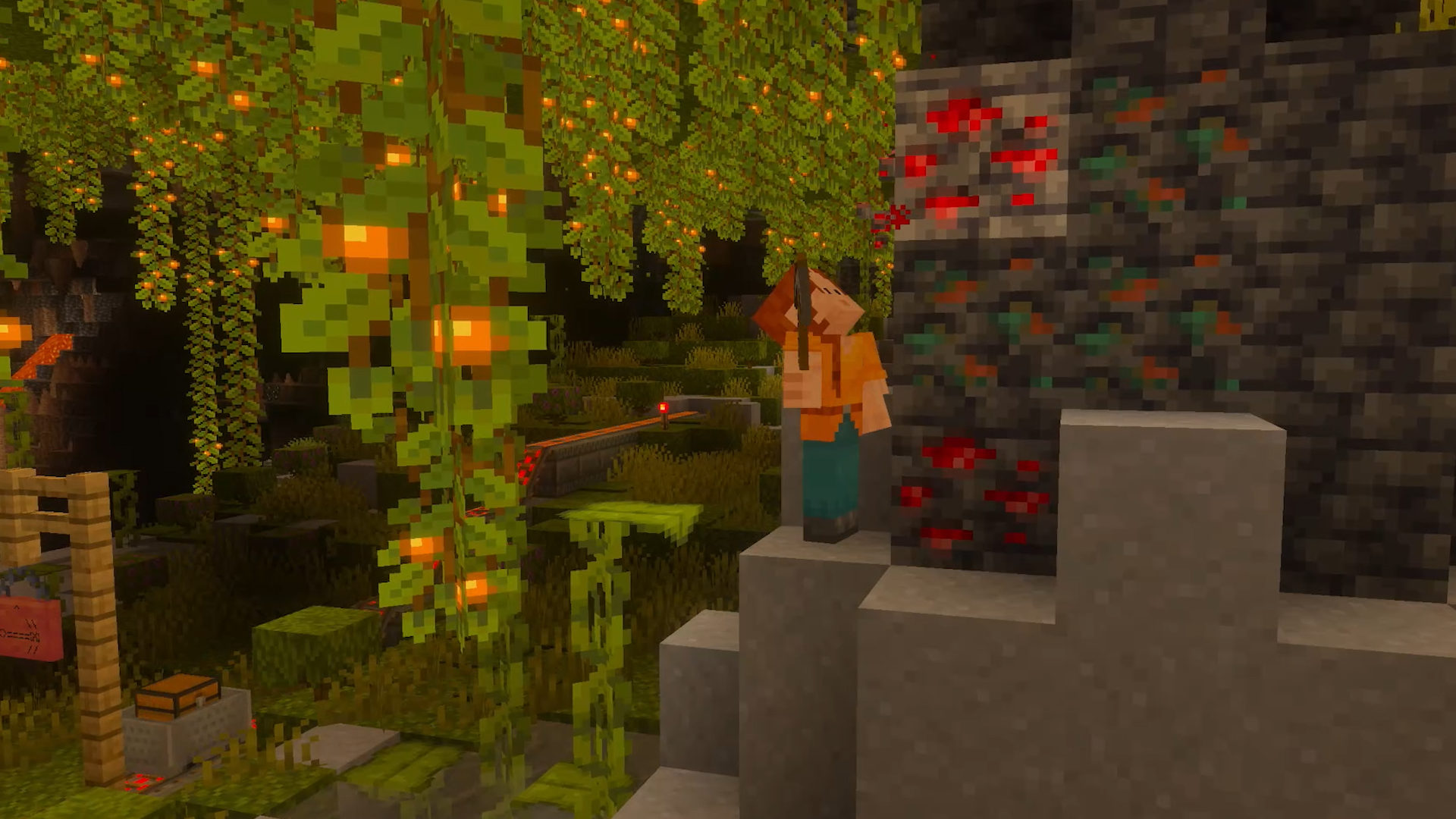 Capture d’écran de Minecraft montrant un personnage en blocs debout dans une grotte luxuriante, près d’un mur de minerais rouges et verts. Des lianes lumineuses, de la végétation dense, des rails et un coffre composent un décor souterrain chaleureux, mêlant exploration et ressources à collecter.