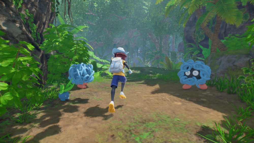 Capture de gameplay d’un jeu d’aventure avec créatures, montrant une jeune exploratrice courant dans une jungle luxuriante. Elle est accompagnée de petites créatures bleues aux formes végétales, dont deux ressemblent à des amas de lianes aux grands yeux.