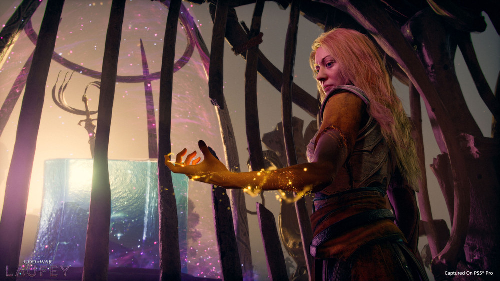 Scène cinématique de jeu vidéo God of War: Valhalla montrant une femme blonde en armure, vue de profil, dans une structure sombre faite de barres de bois. Elle tend la main vers l’avant, entourée de particules dorées magiques qui s’écoulent sur son bras. En arrière-plan, un cube lumineux et un portail cosmique avec des effets stellaires violets et roses. Atmosphère mystique et dramatique, avec mention “Captured on PS5 Pro” en bas à droite.