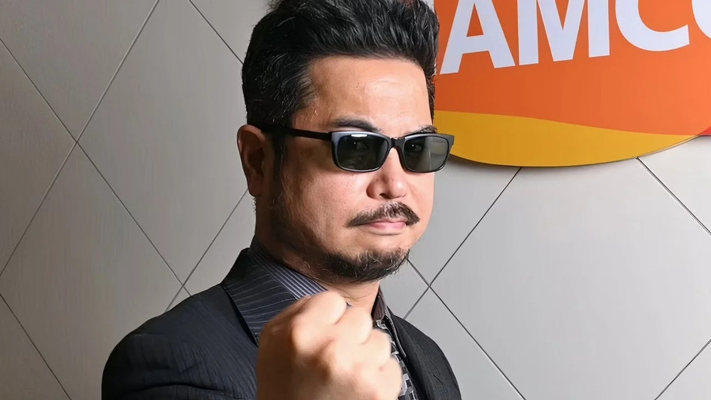 Katsuhiro Harada, producteur emblématique de la série Tekken, pose face à l’objectif en costume sombre rayé, chemise légèrement entrouverte et lunettes de soleil noires. Il affiche une expression sérieuse et déterminée, le poing serré levé vers l’avant dans un geste symbolique d’assurance ou de défi. À l’arrière-plan, on distingue partiellement un logo aux couleurs orange, jaune et blanche appartenant à Bandai Namco, renforçant l’aspect professionnel et lié à l’industrie du jeu vidéo de la scène. Le mur en arrière-plan est décoré de motifs géométriques discrets.