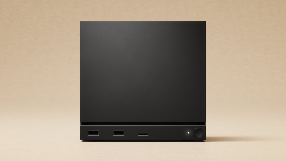 Visuel promotionnel montrant un boîtier informatique noir au design minimaliste, présenté de face sur un fond beige. La façade intègre deux ports USB, un lecteur de carte mémoire, un bouton d’alimentation circulaire accompagné d’un voyant lumineux et une grande partie supérieure lisse aux lignes épurées.