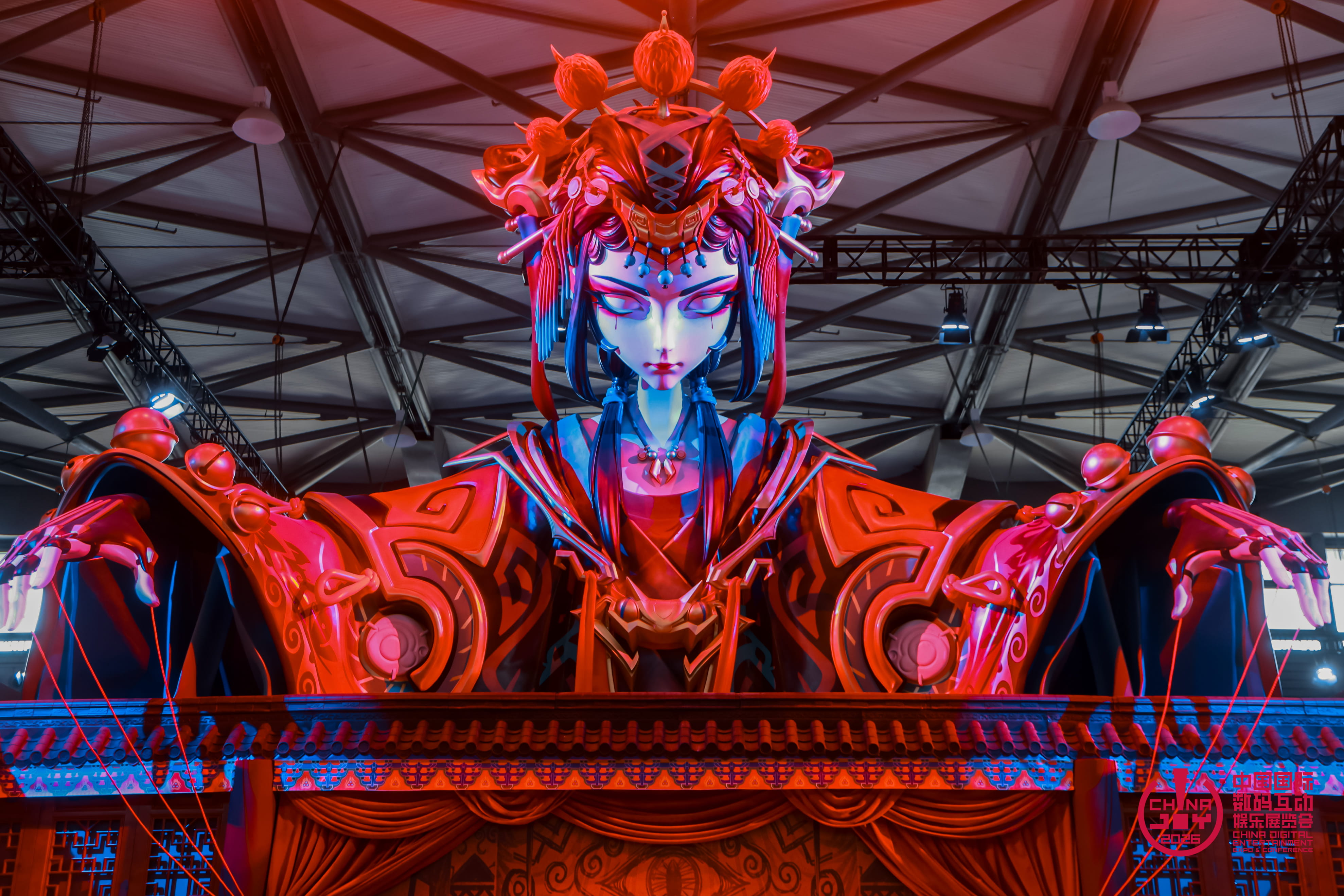 Installation monumentale à ChinaJoy 2026 représentant un personnage féminin stylisé inspiré de l’esthétique chinoise traditionnelle, vêtu d’une tenue rouge et noire avec une imposante coiffe. Éclairée de rouge et de bleu, la figure tient ses bras écartés au-dessus d’un décor évoquant un théâtre traditionnel.