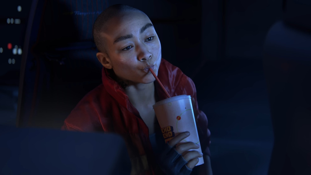 Capture d’écran d’un jeu vidéo montrant un personnage au crâne rasé vêtu d’une veste rouge, buvant avec une paille dans un grand gobelet de boisson. La scène se déroule dans un intérieur sombre éclairé par des lumières bleutées, avec un gros plan mettant en avant son expression détendue et le gobelet tenu à deux mains.