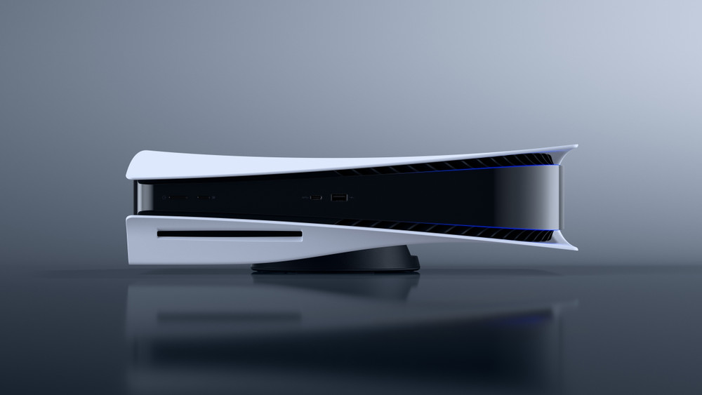 Console PlayStation 5 blanche et noire présentée à l’horizontale sur son socle, vue de face sur un fond gris. Le lecteur de disque est visible à gauche, avec les ports en façade et un éclairage bleu discret le long de la coque.