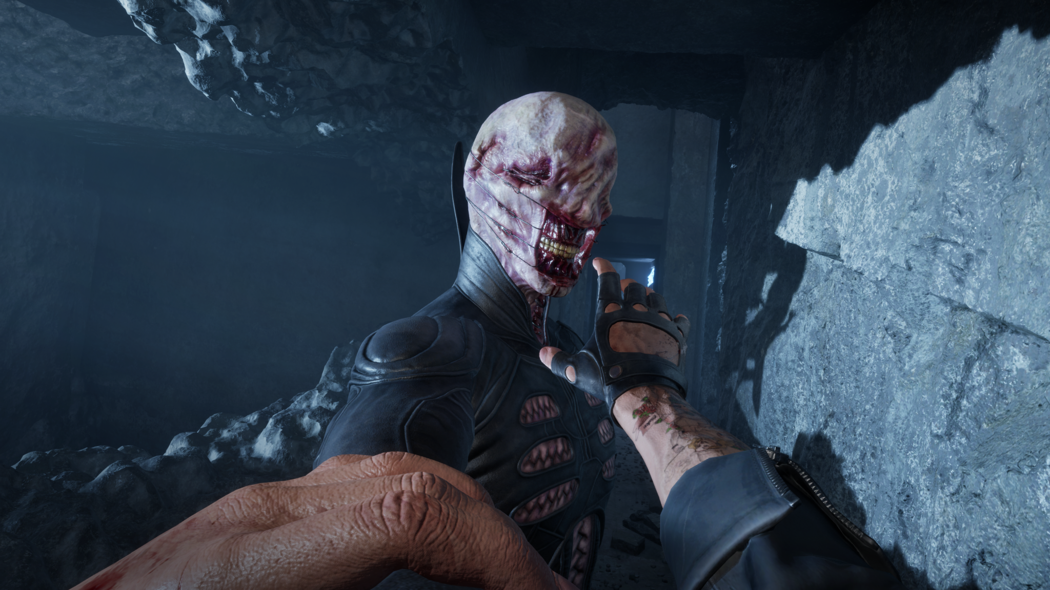 Capture d’un jeu d’horreur à la première personne montrant le joueur aux prises avec une créature humanoïde au visage mutilé et cousu, vêtue d’une combinaison noire, dans un passage rocheux sombre.