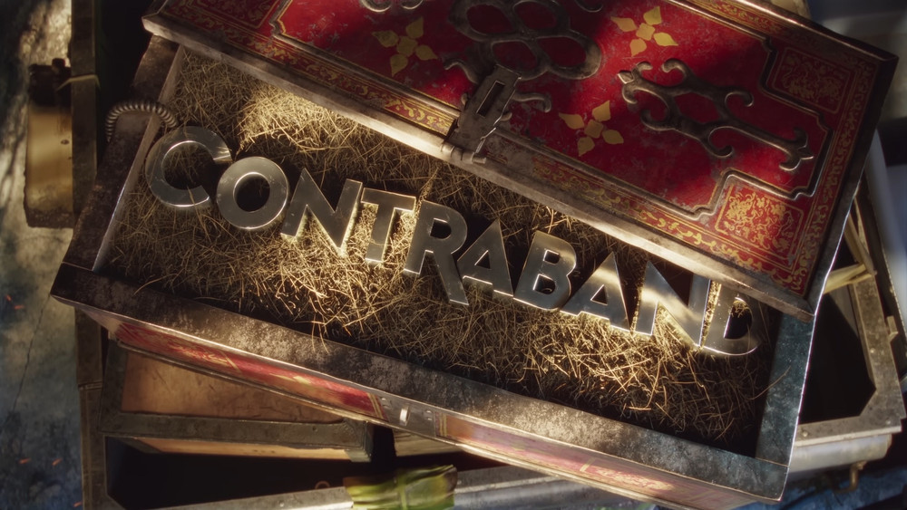 Gros plan sur un coffre ancien entrouvert, au couvercle rouge orné de motifs dorés et de ferronneries noires, laissant apparaître à l’intérieur des lettres métalliques formant le mot « CONTRABAND ». Les lettres reposent sur un lit de paille sèche, éclairées par une lumière chaude qui accentue leur brillance. Le coffre, usé par le temps, est posé sur d’autres caisses en bois et métal, suggérant un contexte d’aventure ou de contrebande. L’image évoque un univers de trésors cachés et de mystère, avec une ambiance à la fois exotique et clandestine.