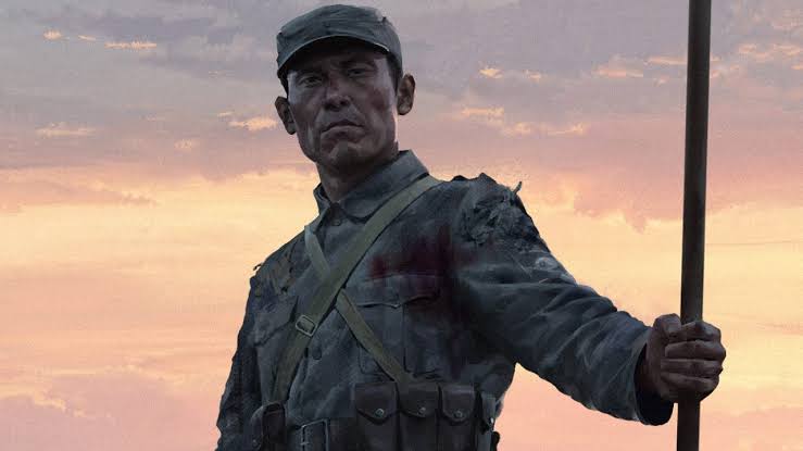 Soldat chinois en uniforme gris, debout au coucher du soleil, tenant une longue hampe verticale dans sa main droite, avec un visage marqué et une tenue usée évoquant un contexte de guerre historique.