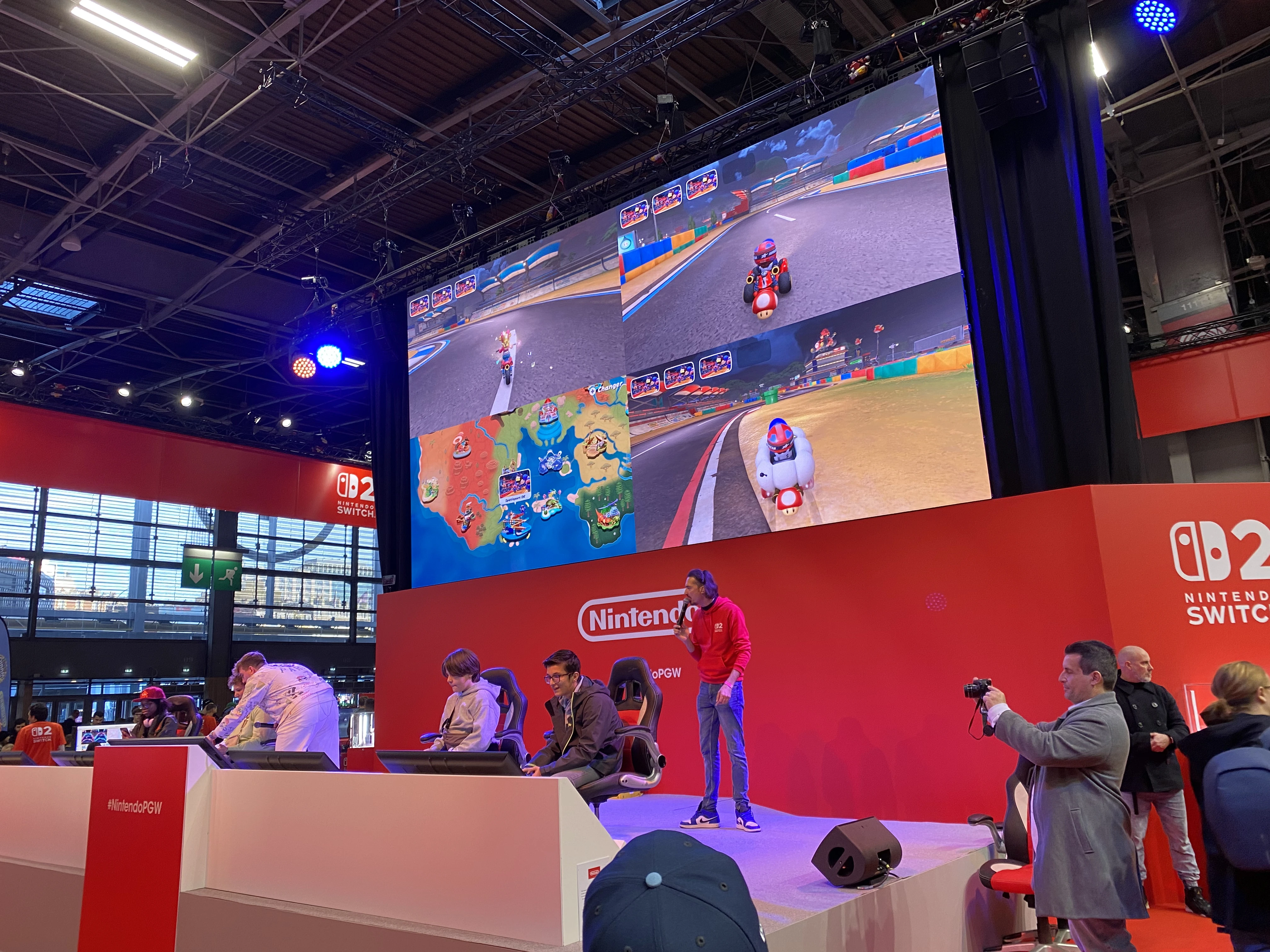 Scène de compétition de Mario Kart 8 Deluxe sur le stand Nintendo à la Paris Games Week 2025. Plusieurs joueurs, assis sur des sièges de gaming alignés devant des écrans, participent à une partie multijoueur projetée en temps réel sur un immense écran au-dessus d’eux. L’affichage montre une vue en écran partagé avec quatre joueurs en pleine course sur différents circuits colorés. Un animateur en sweat rouge Nintendo se tient debout sur la scène, micro à la main, assurant l’ambiance. À droite, un photographe capture la scène tandis que d’autres visiteurs observent. L’ensemble est habillé aux couleurs de la Nintendo Switch 02, avec une scénographie rouge vif, des logos bien visibles et un éclairage dynamique. L’événement met en avant l’esprit festif, compétitif et familial de la marque Nintendo.