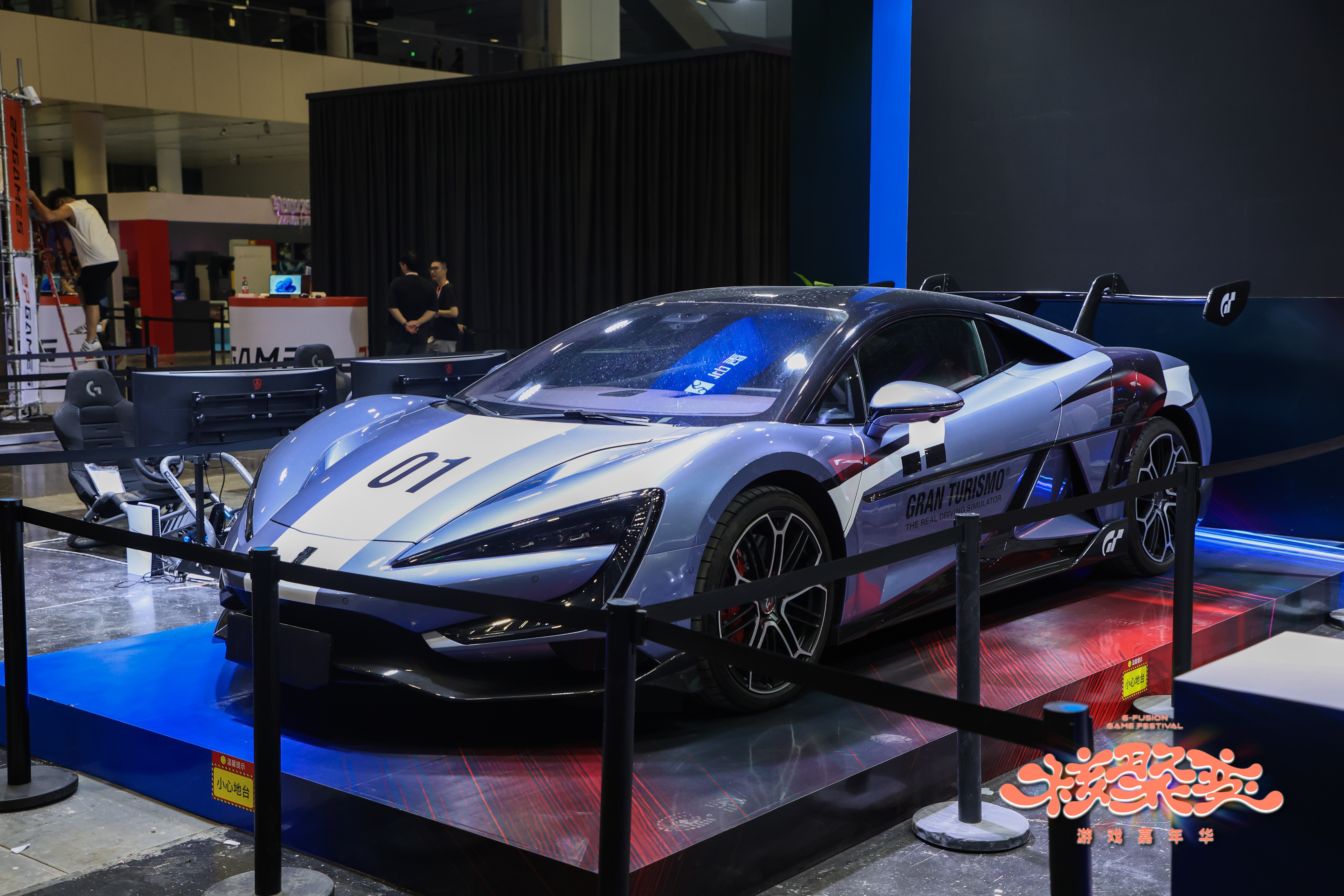 Photographie d’une voiture de sport exposée sur un stand Gran Turismo lors d’un salon du jeu vidéo. Le véhicule gris et blanc porte le numéro 01, le logo Gran Turismo et un grand aileron arrière. Des simulateurs de conduite sont installés derrière la voiture, tandis que des barrières délimitent l’espace d’exposition et que le logo du festival apparaît en bas à droite de l’image.