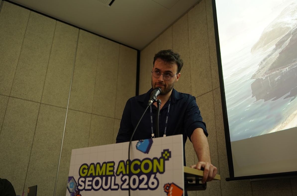 Intervenant au pupitre GAME AiCON Seoul 2026 lors d’une conférence, parlant dans un micro devant un écran projetant une image de paysage côtier. Le panneau du pupitre reprend l’identité visuelle pixel art de l’événement.
