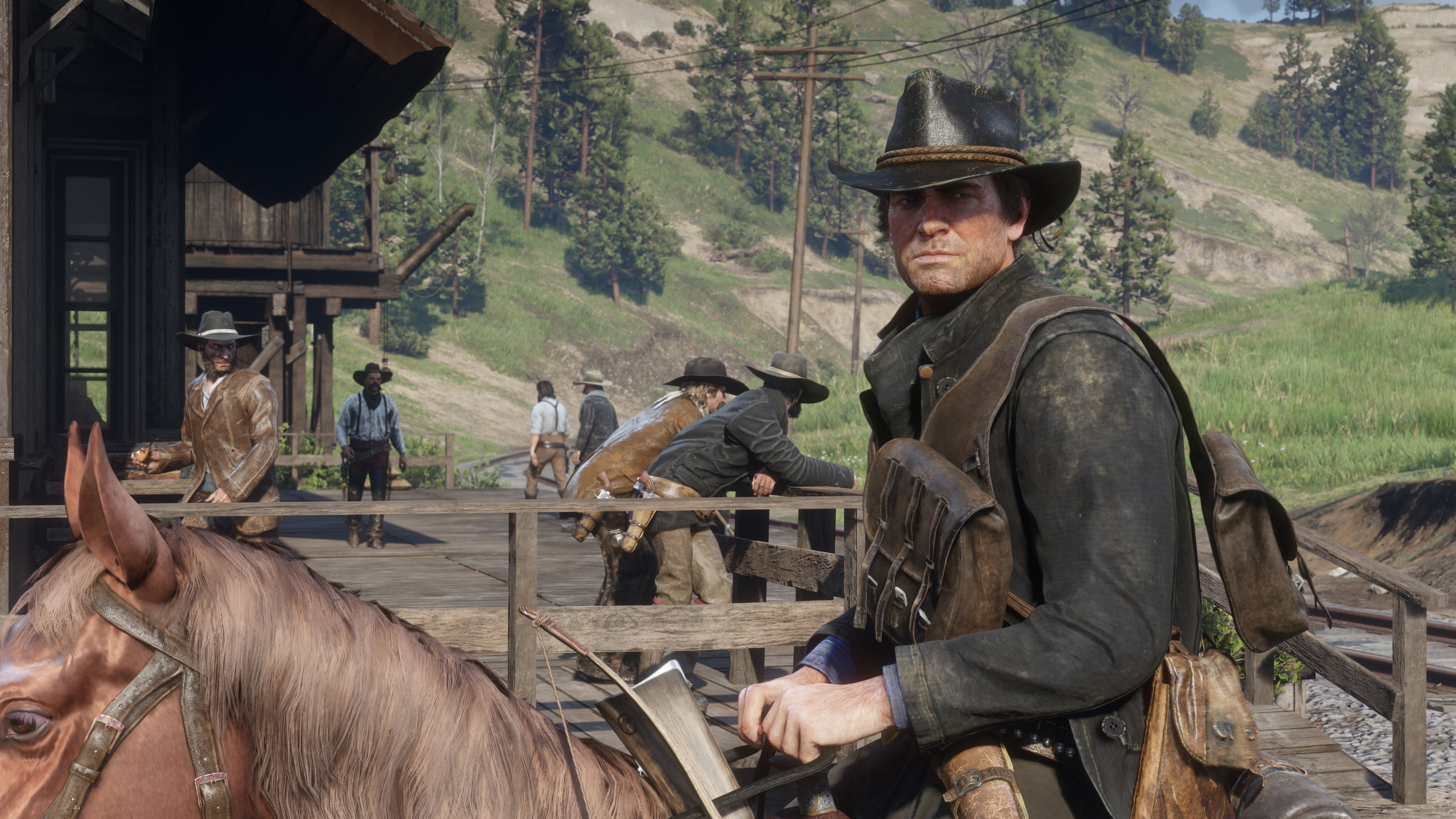 Scène tirée du jeu Red Dead Redemption 2, représentant un homme à cheval, vêtu d’un long manteau en cuir et d’un chapeau de cowboy, regardant directement vers l’objectif avec une expression grave et déterminée. Il s’agit du protagoniste Arthur Morgan, l’un des membres de la bande de Dutch Van der Linde. À l’avant-plan, la tête de son cheval brun occupe une partie de l’image, tandis que l’on distingue dans le dos du cavalier une hache attachée à la selle et une sacoche de voyage. En arrière-plan, plusieurs autres personnages en tenue de l’Ouest sauvage interagissent sur un quai en bois, devant un bâtiment rustique de style western. Certains discutent, d’autres se penchent ou montent la garde. Le décor est composé de collines verdoyantes, de forêts de conifères et de lignes télégraphiques, typiques d’un environnement rural américain du début du XXe siècle. La lumière naturelle, le niveau de détail des textures et la composition réaliste soulignent le soin porté à l’immersion visuelle et historique dans Red Dead Redemption 2, jeu qui mêle narration forte, liberté d’exploration et vie de hors-la-loi dans un monde ouvert vaste et vivant.