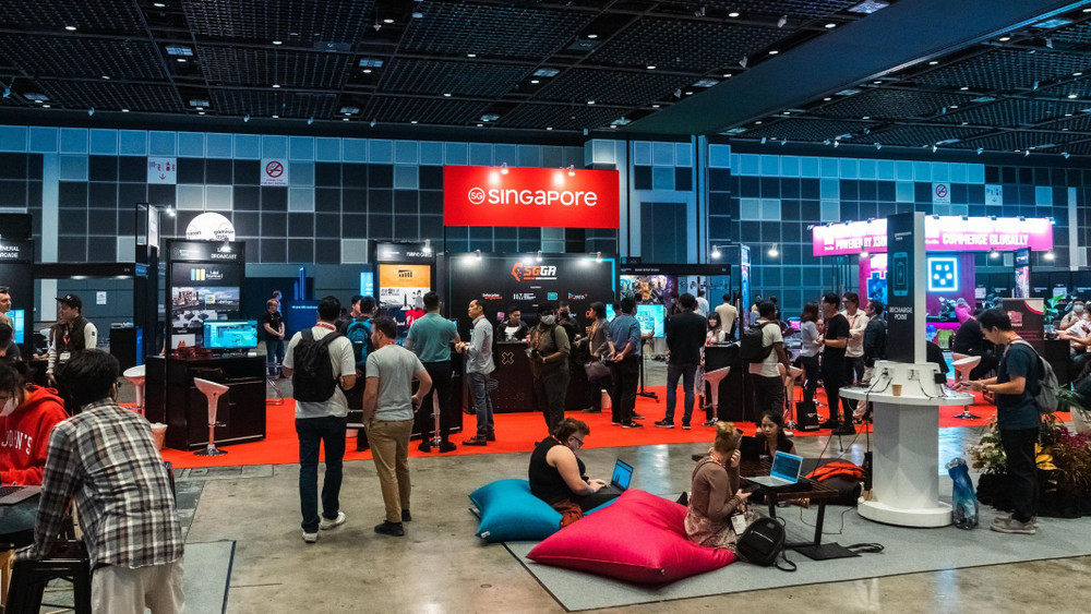 Photographie prise lors d’un salon du jeu vidéo et des technologies interactives, se déroulant dans un grand hall d’exposition moderne. L’espace est animé par de nombreux stands et visiteurs. Au centre, un grand panneau rouge avec le logo officiel de Singapore met en avant la présence de la délégation singapourienne, entourée de plusieurs kiosques présentant des projets, studios et innovations liés au gaming. Des écrans diffusent des démonstrations de jeux vidéo et des présentations techniques, attirant l’attention des participants. On aperçoit des groupes de visiteurs en pleine discussion, certains testant des ordinateurs ou consoles, d’autres observant les présentations. À l’avant-plan, plusieurs personnes sont assises sur des poufs colorés, travaillant sur des ordinateurs portables, reflétant une ambiance à la fois professionnelle et décontractée. L’éclairage est clair et homogène, renforçant l’atmosphère dynamique et collaborative typique des conventions internationales de jeux vidéo comme la Gamescom ou le Tokyo Game Show.