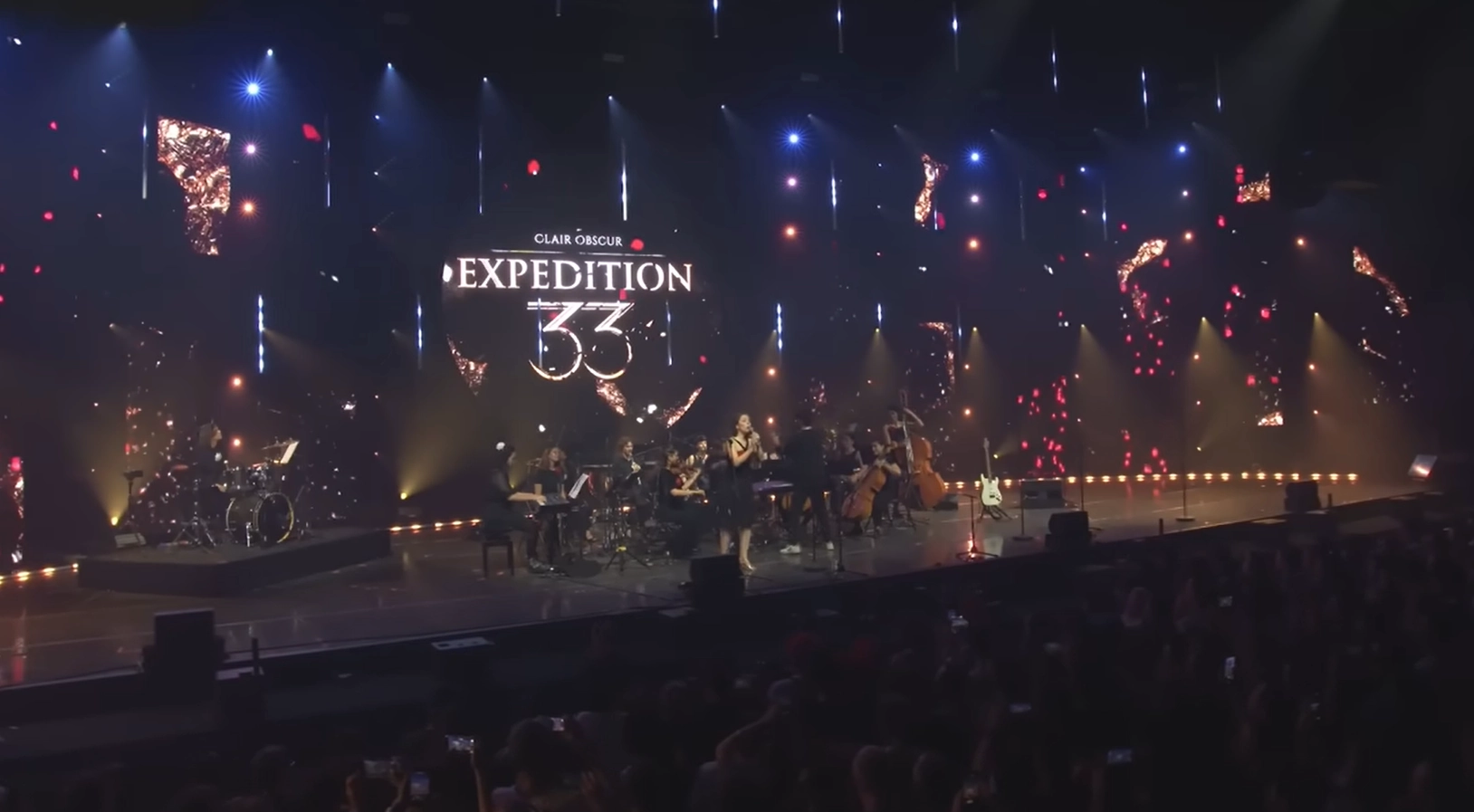 Photo d’un concert en direct intitulé « Clair Obscur : Expédition 33 » lors de la Paris Games Week 2025. La scène est illuminée par un éclairage sophistiqué mêlant projecteurs blancs, jaunes et bleus, ainsi qu’un décor visuel dynamique composé de fragments lumineux rouges et dorés sur un fond sombre. Au centre de l’écran géant derrière les artistes, le titre du spectacle est affiché en grandes lettres : « CLAIR OBSCUR EXPÉDITION 33 ». Sur scène, une chanteuse se tient debout au micro entourée d’un orchestre symphonique avec violons, violoncelles, contrebasses, piano, batterie et guitares. Le public, partiellement visible au premier plan, est plongé dans l’obscurité, certains spectateurs levant leurs téléphones pour filmer la performance. L’ensemble crée une ambiance immersive et cinématographique, renforçant l’aspect narratif et artistique du concert.
