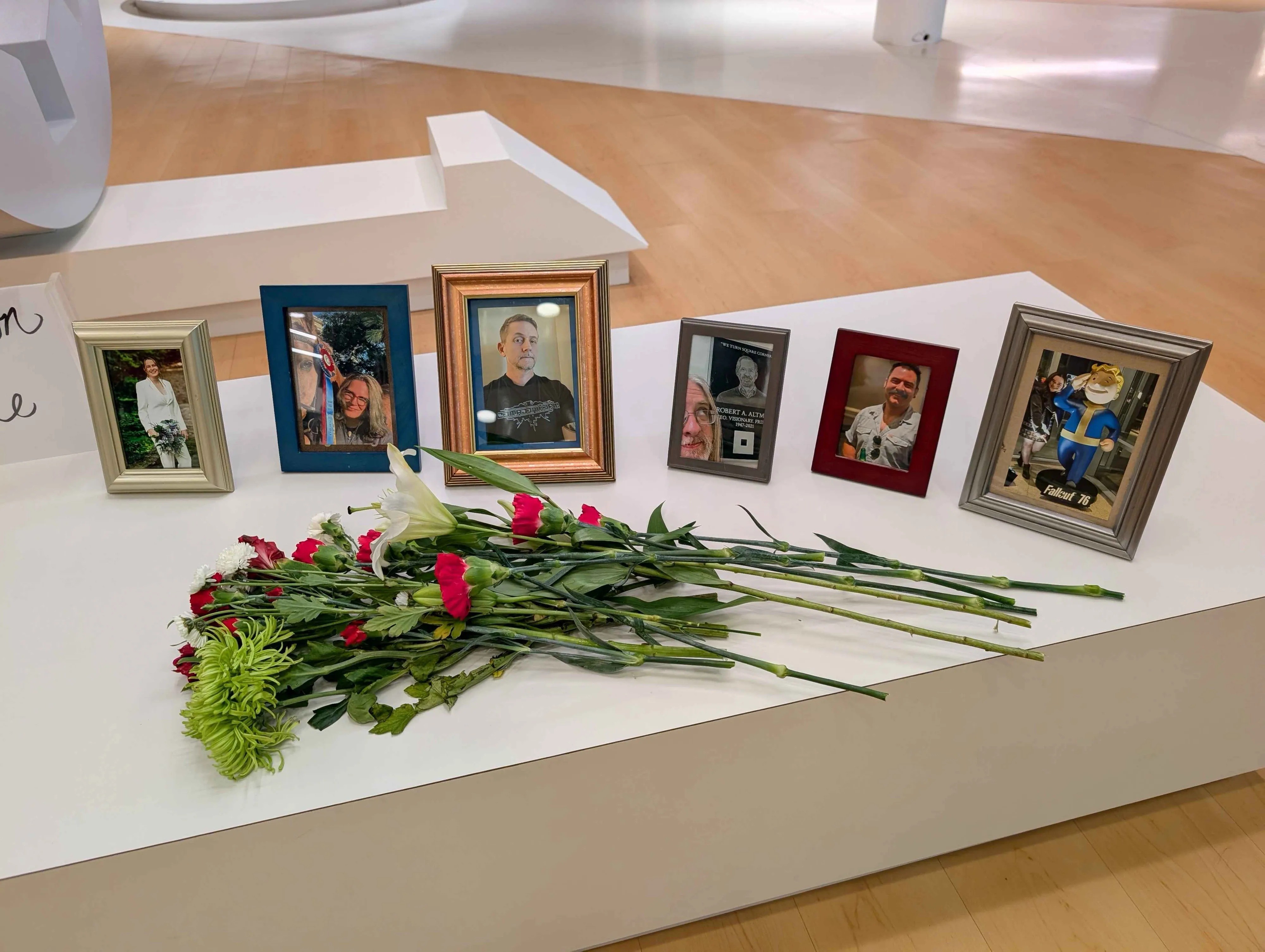 Photographie d’un mémorial installé dans un hall moderne, montrant six photos encadrées de personnes alignées derrière un bouquet de fleurs blanches, rouges et vertes déposé sur un socle blanc. L’un des cadres affiche une image de la mascotte Vault Boy de Fallout 76, tandis qu’une partie du panneau « Celebration of Service » est visible à gauche, évoquant un hommage à des membres de la communauté du jeu vidéo.