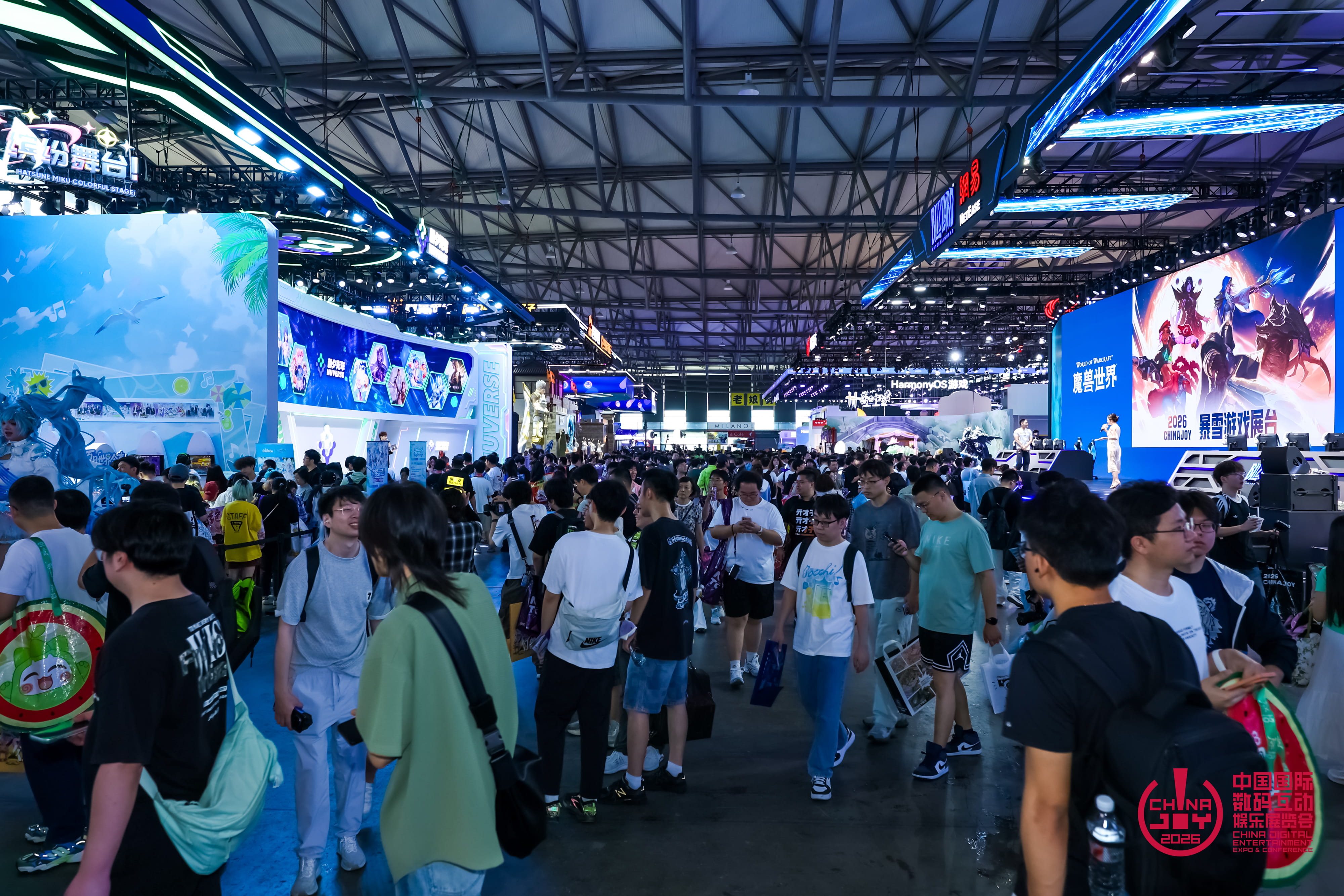 Allée très fréquentée de ChinaJoy 2026, bordée de grands stands de jeux vidéo aux éclairages bleus et d’écrans géants. De nombreux visiteurs circulent entre les espaces d’exposition, tandis qu’une scène à droite présente des visuels de personnages de jeux.