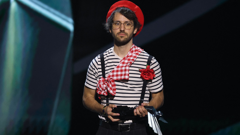 Photographie d’un homme sur scène portant un béret rouge, un t-shirt à rayures, des bretelles et un foulard à carreaux, dans une tenue inspirée d’un mime français. Il tient un trophée dans les mains et affiche une expression sérieuse, avec une grande fleur rouge fixée à sa poitrine et un fond de scène sombre éclairé par des projecteurs.