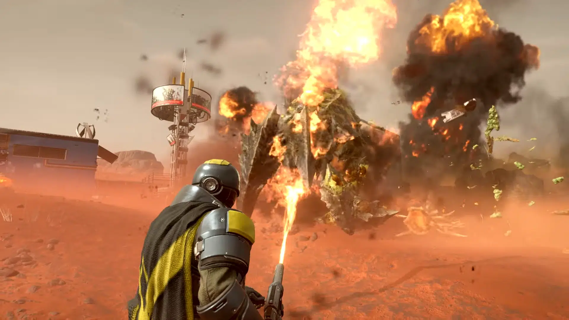 Scène d’action explosive issue du jeu Helldivers II, montrant un soldat futuriste en armure de combat affrontant un gigantesque monstre insectoïde sur un champ de bataille désertique. Au premier plan, le personnage, vu de dos, porte un casque intégral, une cape noire à rayure jaune et une combinaison renforcée, tout en maniant un lance-flammes qui projette un puissant jet de feu vers la créature. Face à lui, une bête massive à la carapace épaisse, couverte d’écailles, est engloutie dans une explosion impressionnante. Des débris, de la fumée noire et des flammes s’élèvent dans le ciel ocre, tandis que des morceaux de la créature sont projetés en l’air. À l’arrière-plan, on distingue un bâtiment bleu, une antenne de communication futuriste et un paysage aride aux formations rocheuses, suggérant une planète lointaine ou colonisée. L’ambiance visuelle est chaotique, intense et cinématographique, représentant parfaitement le style de Helldivers II, un jeu de tir coopératif en vue à la troisième personne où les joueurs doivent survivre à des vagues d’ennemis extraterrestres dans des environnements hostiles.