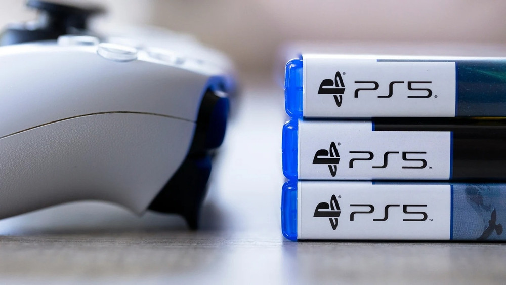 Photographie montrant une manette PlayStation 5 floue au premier plan à gauche et une pile de trois boîtiers de jeux PS5 au premier plan à droite. Les logos PS5 sont clairement visibles sur les tranches blanches des boîtes, tandis que les jaquettes restent partiellement hors champ ou illisibles.