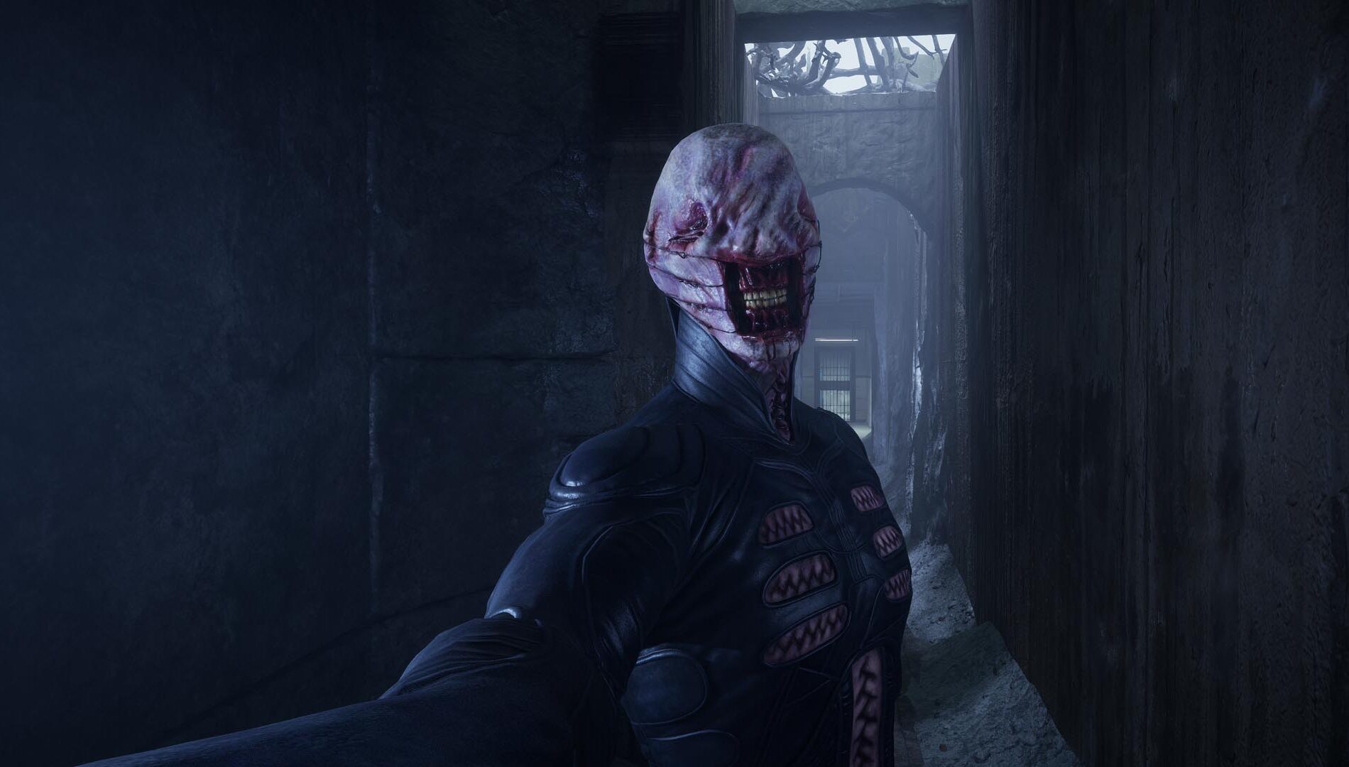 Capture de jeu montrant une créature humanoïde inquiétante dans un couloir sombre, vêtue d’une combinaison noire. Son visage pâle et déformé est dépourvu d’yeux visibles, avec une large bouche laissant apparaître ses dents et plusieurs ouvertures organiques sur le torse.