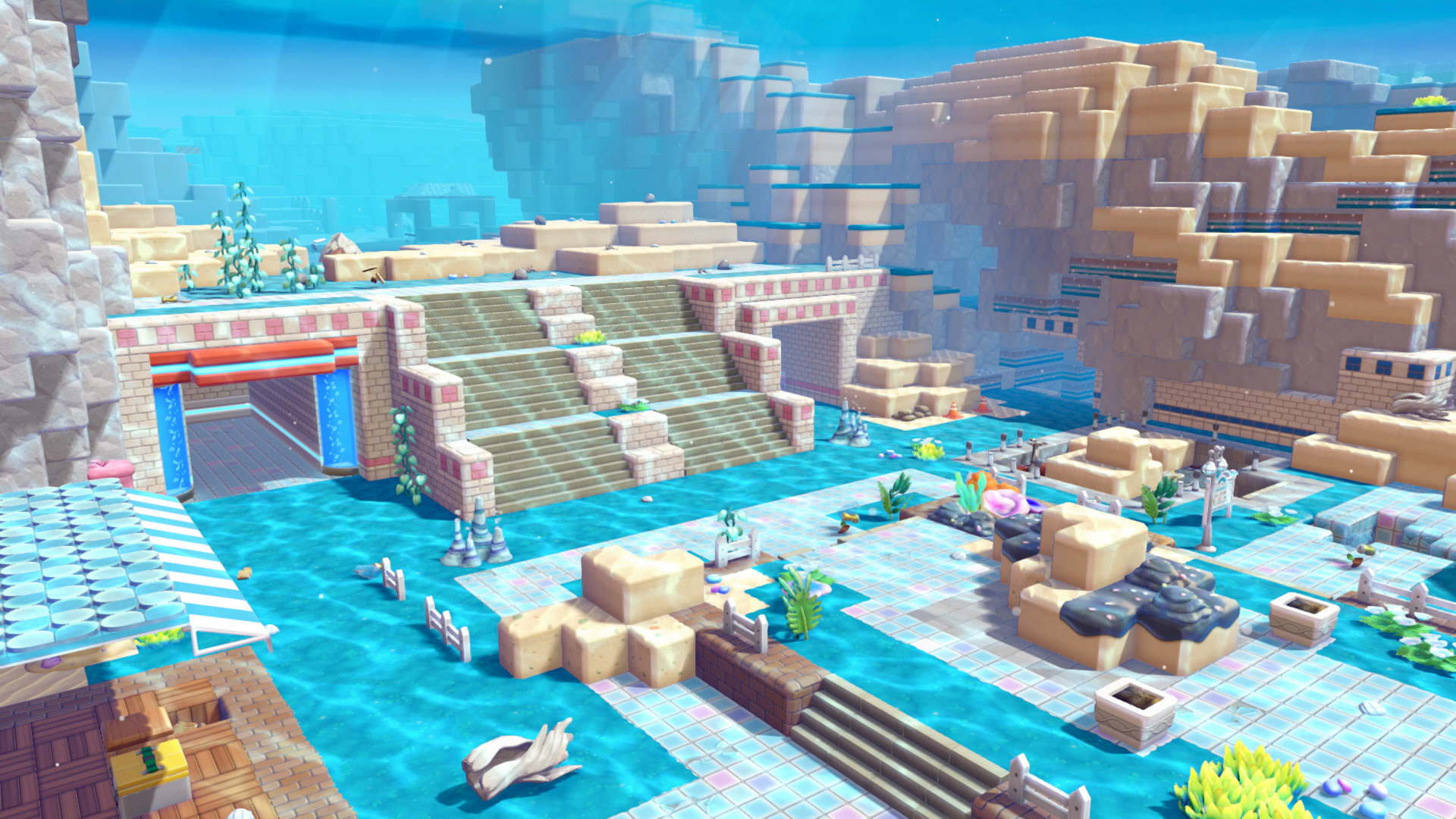 Capture d’écran de Pokémon Pokopia montrant un décor coloré et cubique avec de l’eau turquoise, des escaliers, des plateformes carrelées et des falaises sableuses. L’environnement évoque un village aquatique en construction, décoré de plantes, coquillages, barrières et petits éléments de décor dans une ambiance paisible et lumineuse.