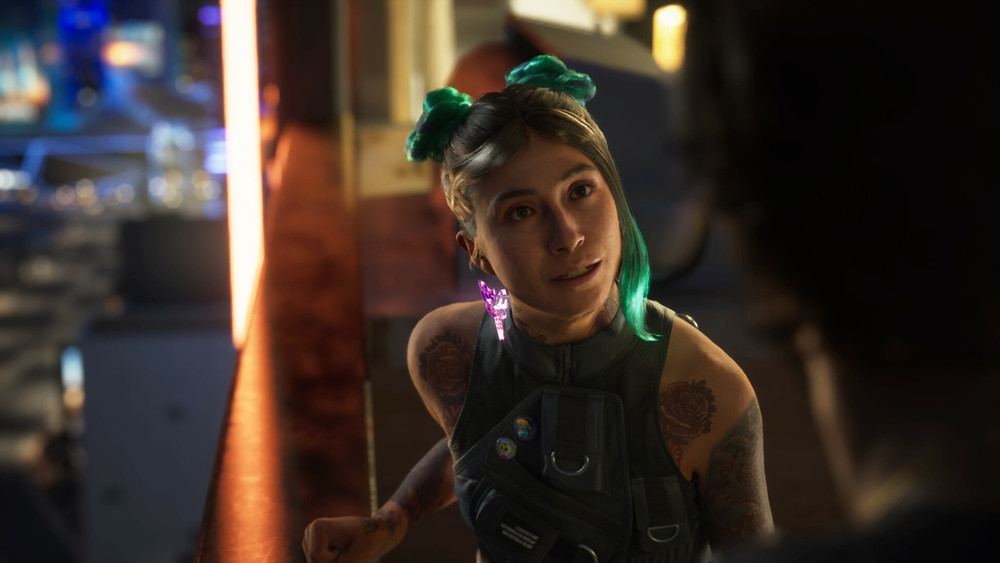 Capture de Cyberpunk 2077 montrant une jeune femme aux cheveux noirs et mèches vertes, coiffée de deux chignons, discutant avec un personnage hors champ dans un bar ou un restaurant futuriste. Elle porte une tenue noire, plusieurs tatouages visibles sur les bras, des badges sur sa poitrine et une boucle d’oreille lumineuse violette, tandis que l’éclairage chaud et les néons créent une ambiance cyberpunk.