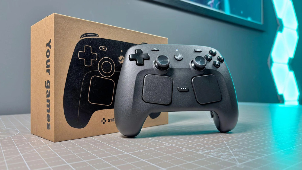 Photo produit d’une manette de jeu vidéo grise de type Steam Deck/Steam Controller posée sur un bureau de découpe, accompagnée de son emballage en carton à l’arrière. Le boîtier affiche une illustration stylisée de la manette et le texte “Your games” sur le côté. La scène est éclairée par une lumière studio avec une ambiance moderne, incluant un éclairage bleu néon à l’arrière-plan, suggérant un produit gaming orienté PC et cloud gaming.