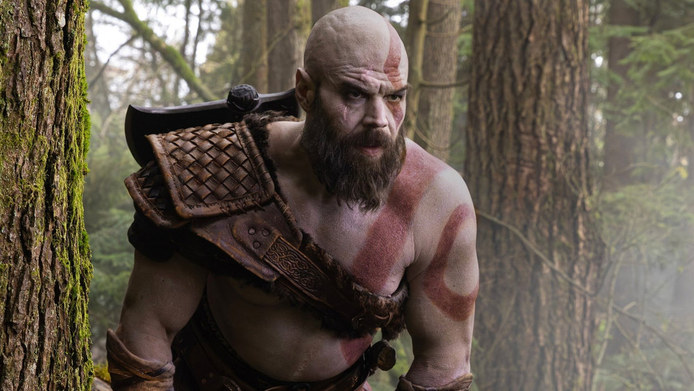 Kratos, guerrier chauve et barbu à la peau pâle marquée d’un tatouage rouge, avance dans une forêt dense. Il porte une armure de cuir sur l’épaule et une grande hache dans le dos, le regard tourné vers l’avant.