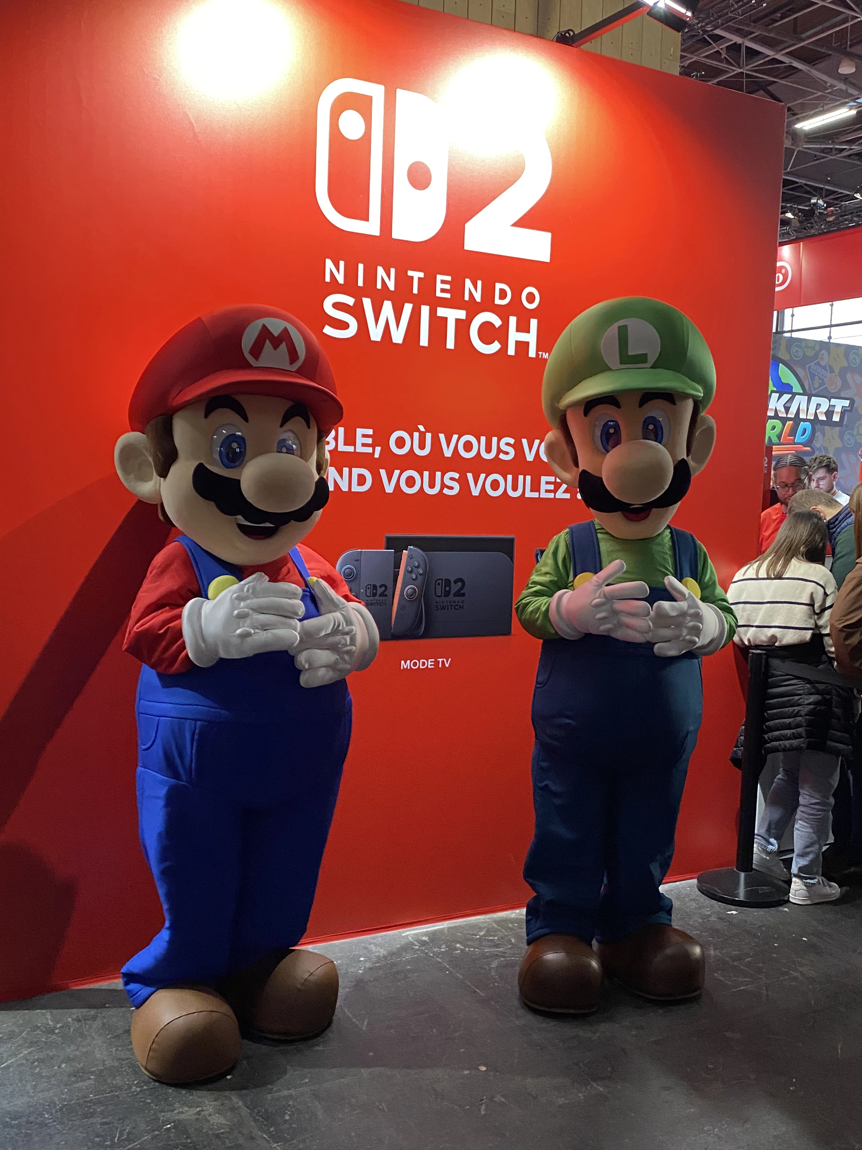 Deux mascottes grandeur nature de Mario et Luigi posent devant un mur rouge décoré aux couleurs de Nintendo Switch, lors de la Paris Games Week 2025. Mario, vêtu de sa salopette bleue, son t-shirt rouge et sa casquette rouge marquée d’un « M » blanc, se tient à gauche, tandis que Luigi, en salopette bleue, t-shirt vert et casquette verte ornée d’un « L » blanc, est à droite. Le fond affiche le logo de la Nintendo Switch ainsi que le nom du nouveau modèle « Switch 02 » accompagné du slogan en français : « Jouez ensemble, où vous voulez, quand vous voulez ». On distingue également un écran avec une image de la console et, à droite de l’image, quelques visiteurs du salon dans une file d’attente devant le stand, avec en arrière-plan une affiche du jeu Mario Kart.