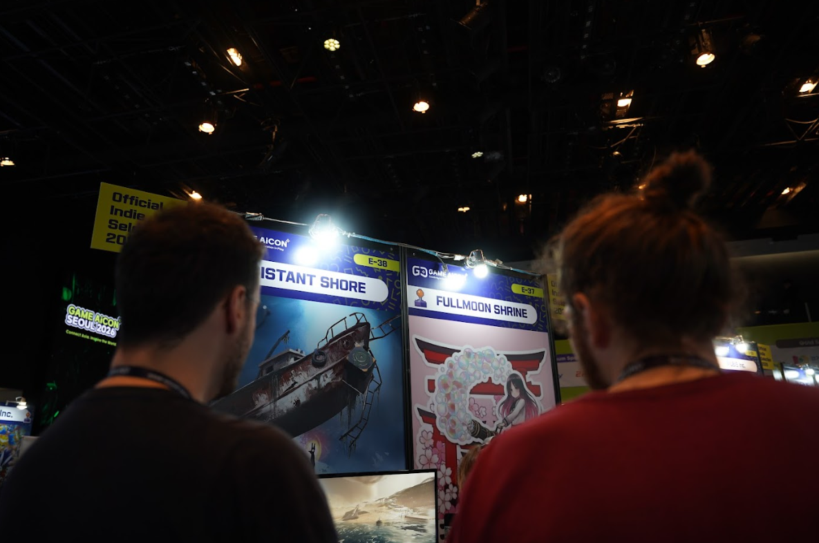 Deux visiteurs vus de dos consultent des stands de jeux indépendants au salon GAME AiCON Seoul 2026. Les panneaux « Distant Shore » et « Fullmoon Shrine » sont visibles sous les éclairages du hall, avec la mention « Official Indie Selection 2026 » en arrière-plan.