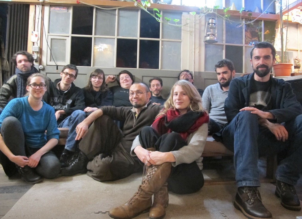 Groupe de dix personnes assises et posant ensemble dans un espace intérieur à l’ambiance chaleureuse, devant une grande fenêtre à carreaux. Les membres, hommes et femmes, sont assis au sol ou sur un canapé bas, souriant ou adoptant une expression détendue. L’environnement comporte des éléments décoratifs comme des plantes suspendues et une lanterne, suggérant un cadre créatif ou associatif, typique d'un studio indépendant de développement de jeux vidéo.