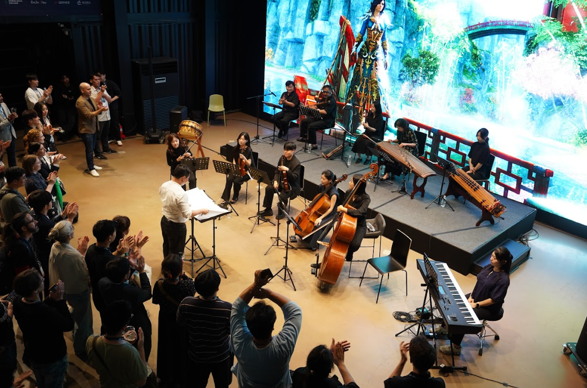 Concert live lors du GAME AiCON Seoul 2026, avec un ensemble de musiciens jouant cordes, piano et instruments traditionnels devant un grand écran affichant un décor fantasy. Le public debout applaudit autour de la scène.