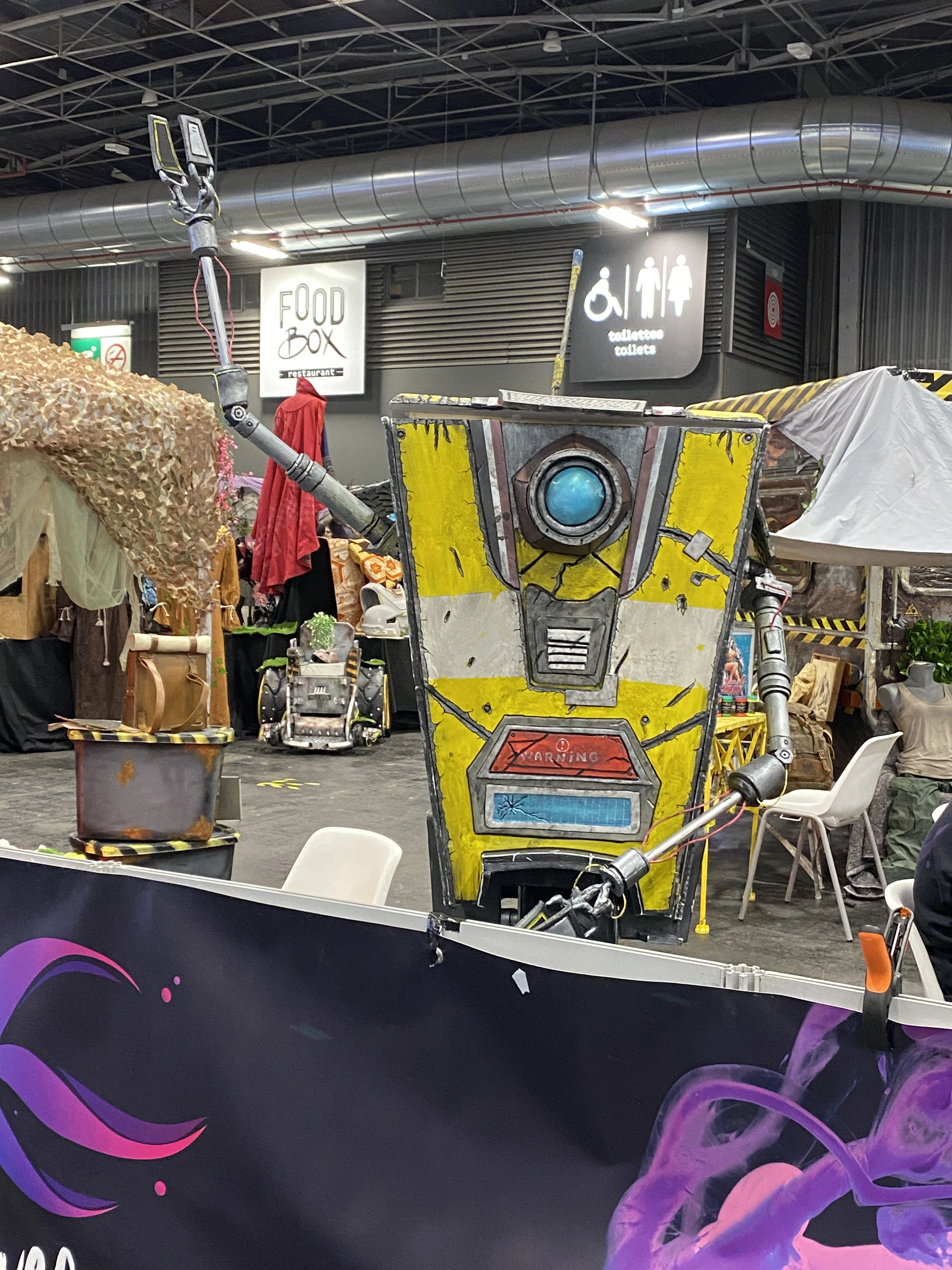 Représentation grandeur nature de Claptrap, le célèbre robot jaune et gris de la franchise Borderlands, exposé à la Paris Games Week 2025. Le personnage, avec son œil caméra lumineux au centre et ses deux bras mécaniques levés, semble saluer ou interagir avec le public. Il est installé dans un espace décoré de manière post-apocalyptique, avec des éléments de décor évoquant l’univers de Borderlands : bidons rouillés, tissus usés, et ambiance industrielle. En arrière-plan, on distingue des stands de restauration avec les enseignes « Food Box » et « Toilets », ainsi que des visiteurs assis sur des chaises blanches. L’avant-plan est occupé par une bannière aux motifs violets et noirs.