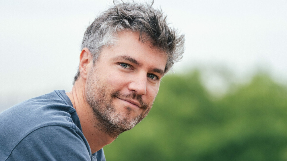 Portrait d’un homme adulte en extérieur, regardant la caméra avec un léger sourire. Il a les cheveux courts et ébouriffés, mêlant du gris et du brun, ainsi qu’une barbe de quelques jours bien entretenue. Ses yeux clairs contrastent avec la douceur de son expression détendue. Il porte un t-shirt bleu-gris simple, et l’arrière-plan flou suggère un cadre naturel, avec des arbres verts en second plan sous un ciel légèrement voilé. La lumière est douce et diffuse, renforçant l’atmosphère paisible et naturelle de la scène. Ce type d’image pourrait être utilisé pour illustrer une interview de développeur de jeu vidéo, un créateur indépendant ou une figure du milieu vidéoludique dans un contexte informel.