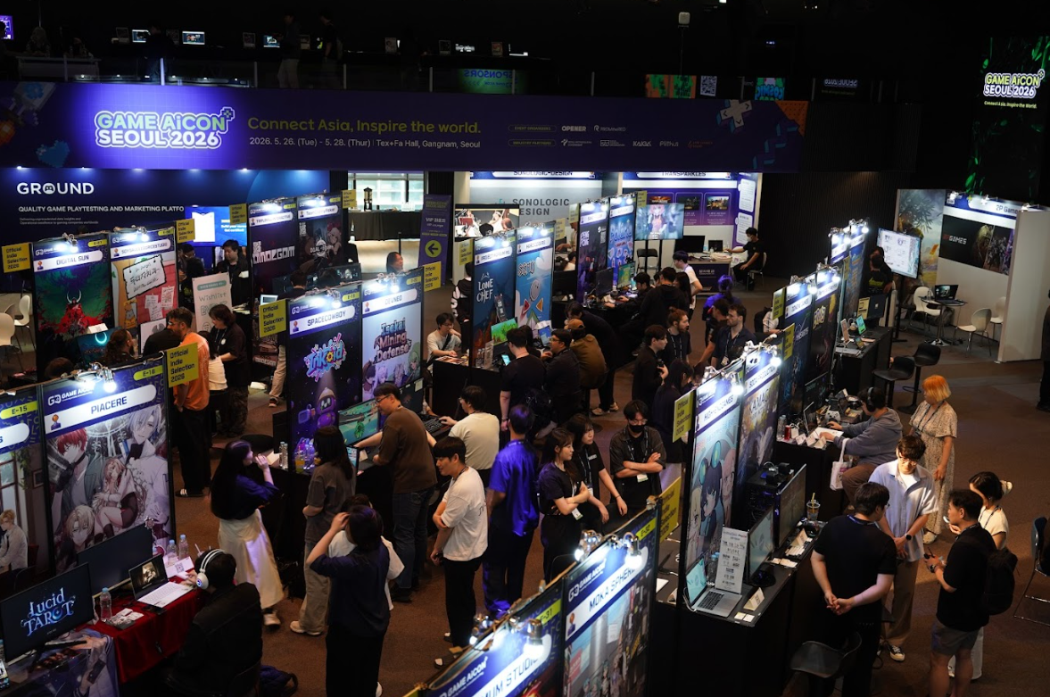 Vue en hauteur du salon GAME AiCON Seoul 2026, avec de nombreux visiteurs circulant entre des stands de jeux indépendants éclairés. Une grande bannière annonce l’événement et son slogan « Connect Asia, Inspire the world », du 26 au 28 mai 2026 au TextFa Hall, Gangnam, Séoul.